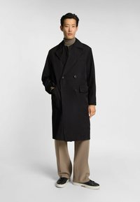 Czarny podwójnie zapięty płaszcz z szerokimi klapami, bocznymi kieszeniami i detalami z guzikami, noszony na beżowym golfie i beżowych spodniach.