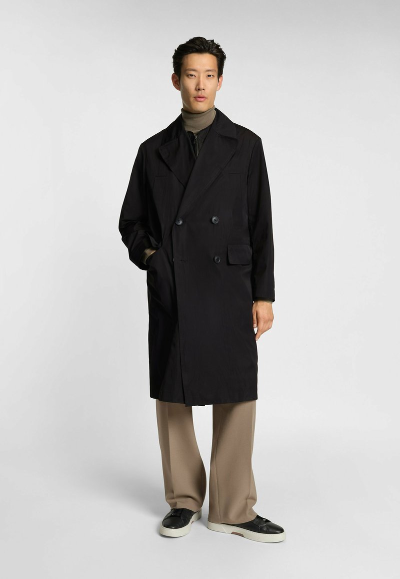 Czarny podwójnie zapięty płaszcz z szerokimi klapami, bocznymi kieszeniami i detalami z guzikami, noszony na beżowym golfie i beżowych spodniach.