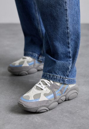 Osoba ubrana w niebieskie dżinsy i szare tenisówki z niebieskimi i beżowymi siateczkowymi detalami, stojąca na gładkiej szarej podłodze.