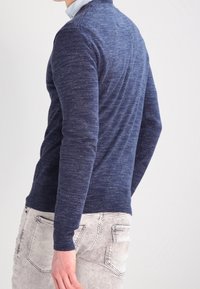 Pull en tricot bleu marine à col rond et manches longues, présentant une texture subtile et une coupe ajustée, porté avec un jean de couleur claire.
