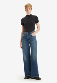 Wijdvallende blauwe denim jeans met een hoge taille, lichte vervaging en opvallende zijzakken. Gecombineerd met een zwart shirt met korte mouwen.