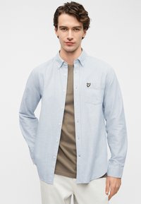 Φωτεινό γαλάζιο πουκάμισο με ρίγες και κουμπιά, με τσέπη στο στήθος που φέρει ένα μικρό κεντητό λογότυπο στην αριστερή πλευρά, φορεμένο πάνω από ένα καφέ T-shirt.