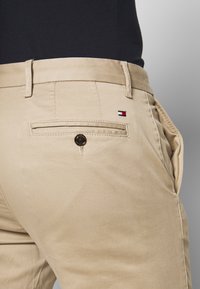 Chinos beige con una texture liscia, dotati di una tasca sul retro con un bottone e un piccolo logo in rosso, bianco e blu.