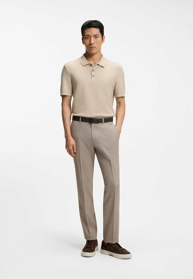 Homme portant un polo beige à manches courtes, un pantalon marron clair avec ceinture noire, et des baskets marron foncé, debout devant un fond blanc uni.