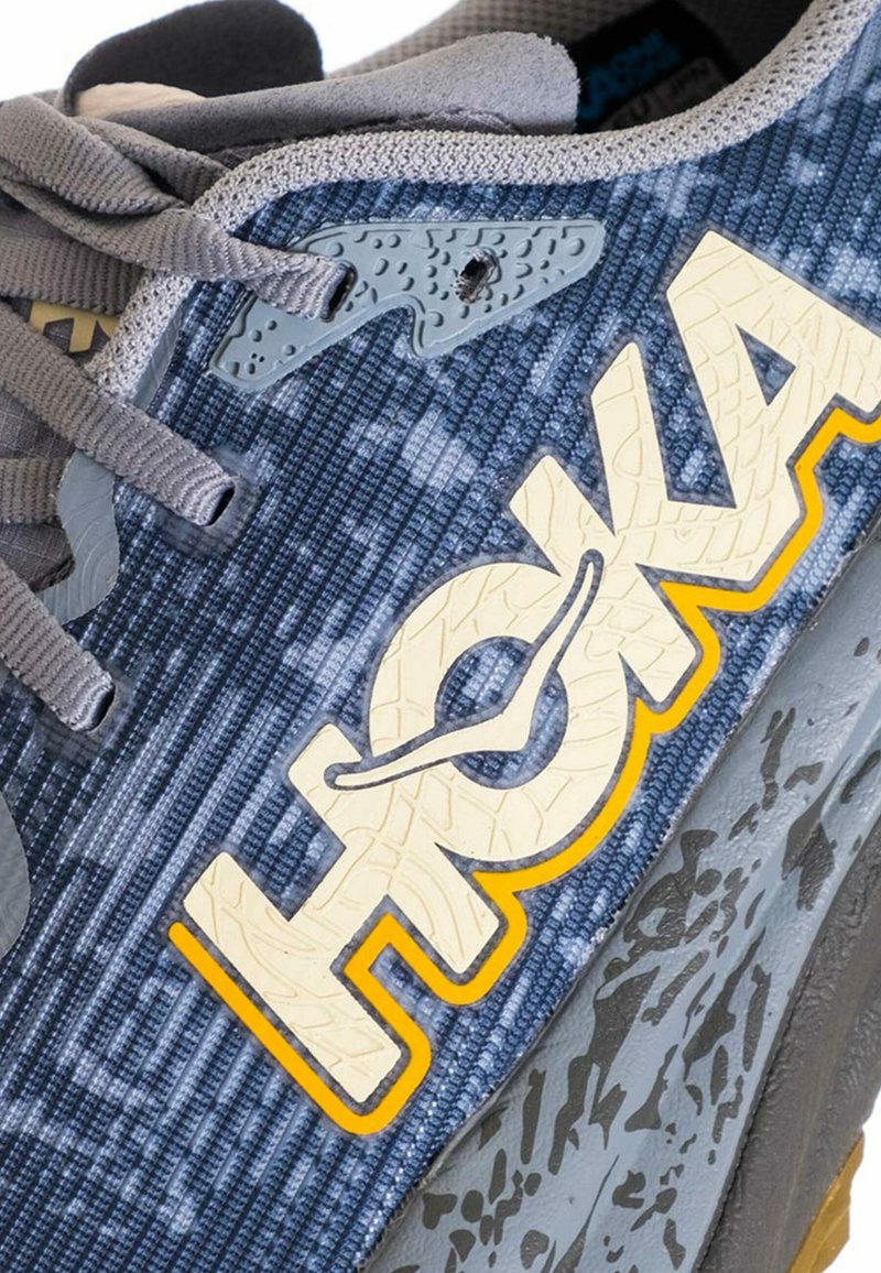 Primo piano di una sneaker grigia e blu con lacci grigi e un grande logo "HOKA" testurizzato color crema e giallo sul lato.