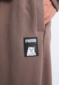 Brun stof med ribbet tekstur, der har en sort label. Labelen viser en tegneseriehvid kat og ordet "PUMA" over den.