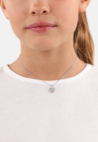 Collana in argento con pendente a forma di cuore caratterizzato da accenti multicolori e lettere incise, indossata su una camicia bianca.