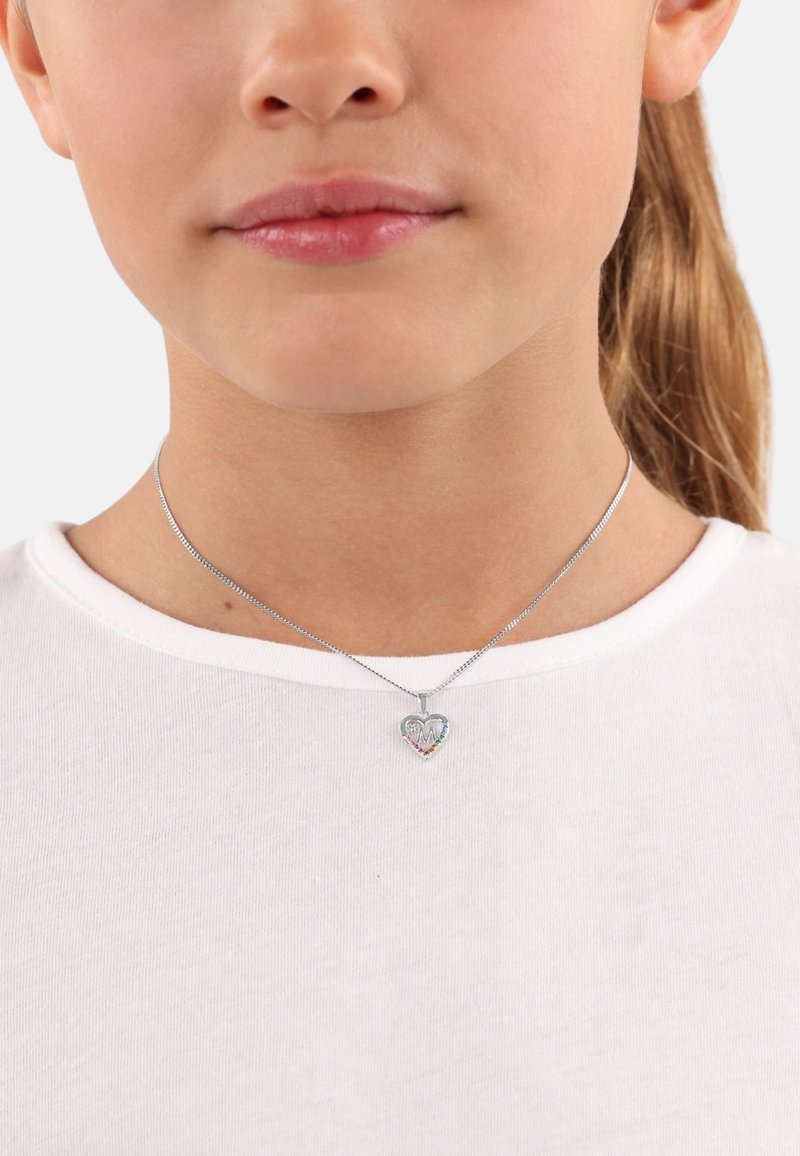 Collana in argento con pendente a forma di cuore caratterizzato da accenti multicolori e lettere incise, indossata su una camicia bianca.