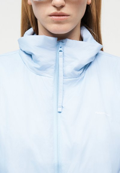 Γυναίκα που φοράει ανοιχτό μπλε ζακέτα με φερμουάρ και ψηλό γιακά που καλύπτει εν μέρει τον λαιμό, με λευκό κείμενο στο στήθος.