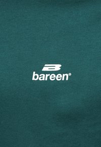 Έντονο λευκό λογότυπο "B" και κείμενο "bareen" στο κέντρο πάνω σε φόντο πετρόλ ύφασμα, πιθανώς μέρος ενός ρούχου.