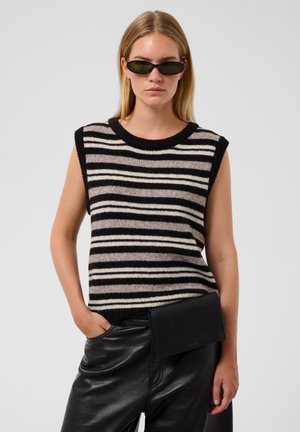 Femme portant des lunettes de soleil noires, un pull sans manches rayé noir, blanc et gris, et un pantalon en cuir noir avec une pochette noire.