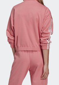 Rosa långärmad jacka i mjukt tyg, med hög krage, elastiska muddar och vita ränder längs ärmarna.