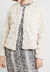 Giacca in pelliccia ecologica color crema con texture trapuntata, caratterizzata da una scollatura rotonda e bottoni a pressione. Indossata sopra un vestito con stampa a motivo serpente.