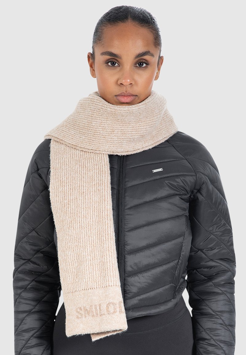 Beige ribbet strikskrave med "SMILE" vævet i enden, båret over en sort polstret jakke. Blød tekstur, medium bredde og lang længde.