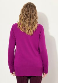 Lila stickad tröja med långa ärmar, ribbad kant och sidofickor, visad bakifrån. Slät textur med en avslappnad passform.