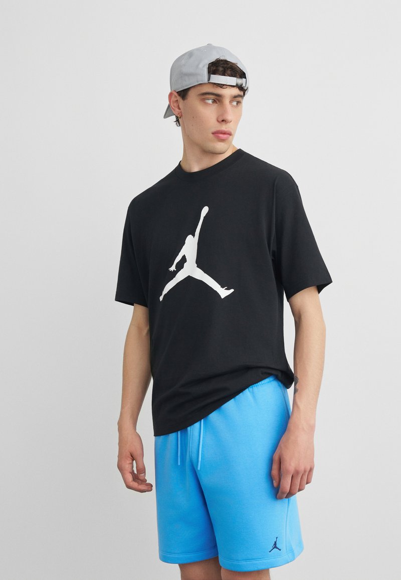 Νέος άνδρας με γκρι καπέλο φοράγμενο ανάποδα, μαύρο μπλουζάκι με λευκή σιλουέτα αλμάτων μπάσκετ και ανοιχτό μπλε σορτς, στέκεται μπροστά σε γκρι φόντο.