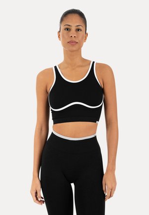 Kobieta stojąca zwrócona twarzą do przodu, ubrana w czarny krótki top z białą lamówką i dopasowane czarne legginsy z wysokim stanem na jednolitym tle.