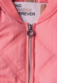 Ljusrosa vadderad jacka med silverfärgad dragkedja, ribbad krage och etikett med texten "HITTA PARADISET FÖR ALLTID".