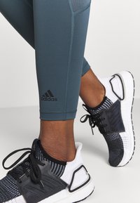 Turkusowe legginsy z czarnym logo Adidas, zestawione z czarno-białymi sneakersami z teksturowanej siateczki i sznurówkami.
