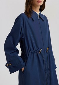 Mornarsko plava, lagana jakna sa patentnim zatvaračem sprijeda, pojasom s vezicom i dugmadima na rukavima. Sadrži dva bočna džepa i klasični ovratnik.