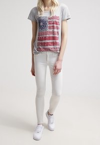 Gray short-sleeve t-shirt with a red and blue American flag graphic, paired with white skinny jeans and white sneakers.