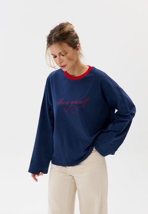 Vrouw met licht haar in een knot draagt een navy sweater met rode tekst "choose yourself", beige broek en kijkt naar beneden tegen een effen achtergrond.