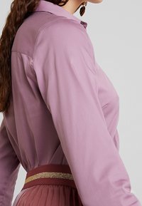 Donna in una camicia a maniche lunghe color malva infilata in una gonna plissettata con una larga cintura bordeaux caratterizzata da una striscia dorata orizzontale.