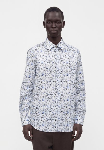 Homme portant une chemise blanche à manches longues boutonnée avec un petit motif floral bleu et noir, debout devant un fond uni.