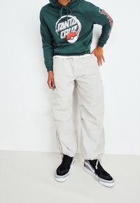 Persona vistiendo una sudadera verde de Santa Cruz con un diseño de Poké Ball, pantalones cargo beige y zapatillas negras de caña alta, de pie contra un fondo blanco.