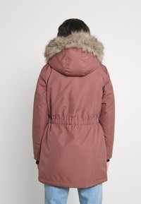 Parka marron avec capuche amovible doublée de fourrure, taille ajustable par cordon, et manches longues, fabriquée en matière lisse et résistante.