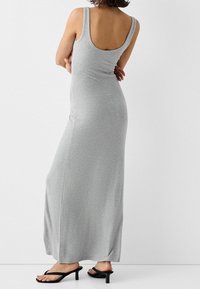 Robe maxi grise, sans manches, près du corps, en tissu doux, avec un dos plongeant et une coupe droite. Portée avec des tongs noires.