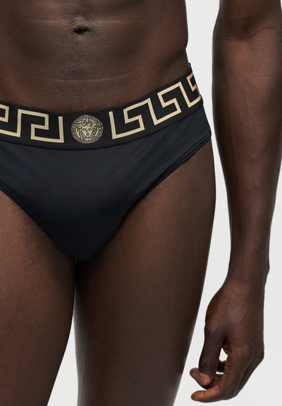 Homme portant un slip noir avec motif de clé grec doré et ceinture à logo tête de Méduse, torse et haut des cuisses visibles.