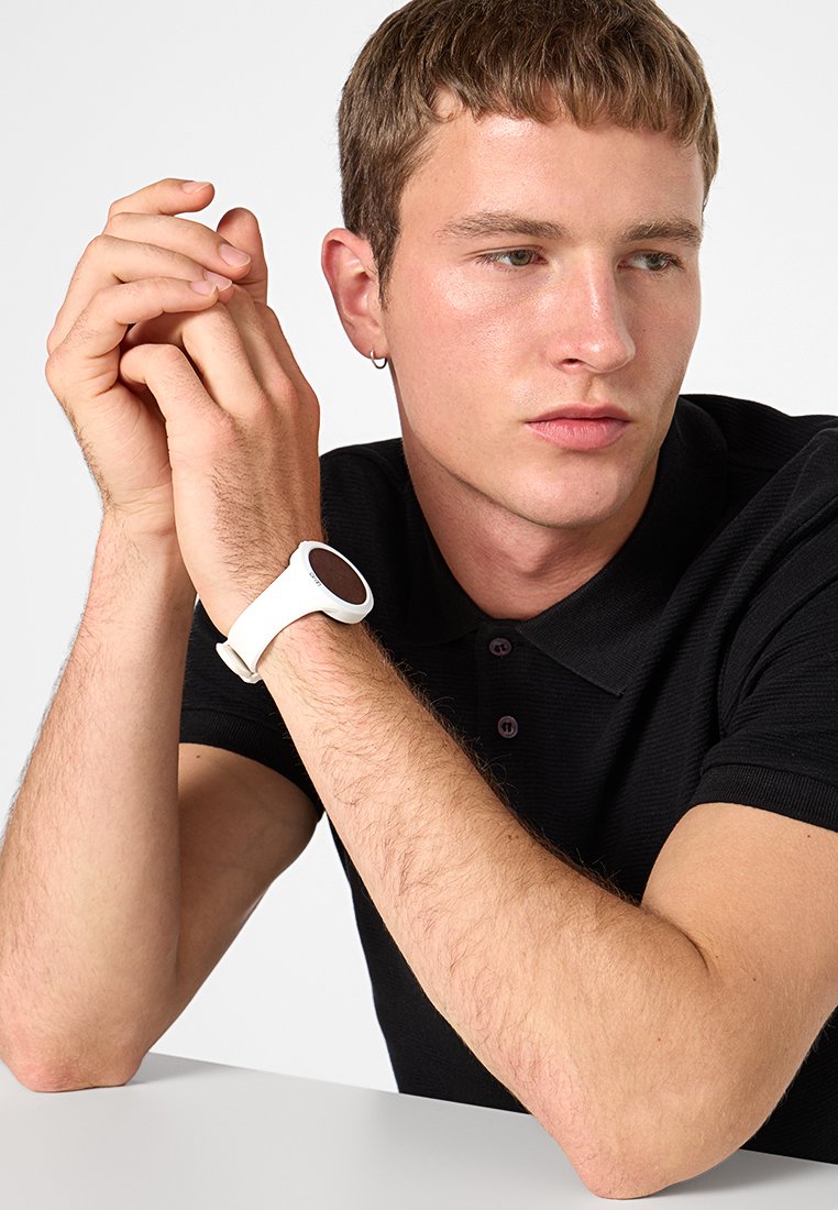 Montre blanche avec un cadran rond et un bracelet lisse. Portée au poignet, avec une zone d'affichage sombre. S'associe avec un polo noir.