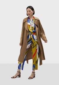 Hnedý trench coat cez viacfarebný odev s živými vzormi, s modrými, žltými, červenými a zelenými akcentmi. Čierne sandále na vysokom opätku.