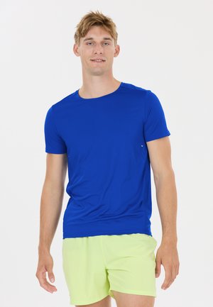 Jonge man staand, gekleed in een felblauw shirt met korte mouwen en lichtgele korte broek tegen een effen witte achtergrond.