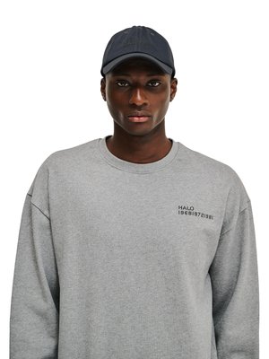 Młody mężczyzna w ciemnej czapce i jasnoszarej bluzie z nadrukiem „HALO 196819721981” na piersi, zwrócony przodem na białym tle.