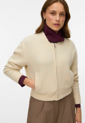 Veste bomber tricotée à zip crème avec des poignets côtelés, des poches latérales et une coupe décontractée, superposée à un col roulé violet foncé.