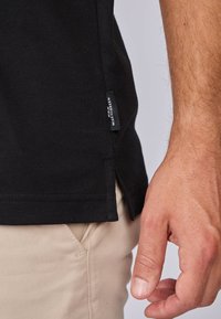 T-shirt en coton noir avec fente sur le côté, présentant une petite étiquette sur l'ourlet. Main reposant sur un pantalon beige clair. Vue en gros plan.