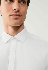 Chemise blanche à boutons avec un tissu texturé. Caractéristiques : col classique, cinq boutons et un design épuré, avec une coupe ajustée autour du cou.