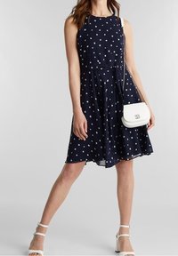 Mörkblå klänning med prickar, utformad med en åtsittande överdel och en utsvängd kjol. Har en midjeband, är ärmlös och en vit axelremsväska.