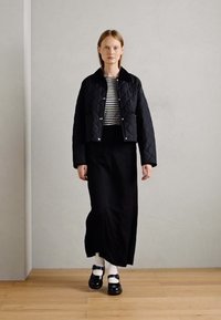 Quilted black jacket with a collar, striped shirt underneath, and a long black skirt. Worn with black shoes and white socks. Wood floor backdrop.