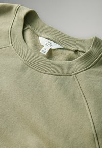 Olivengrüner Sweatshirt mit geripptem Rundhalsausschnitt, weicher Textur und Raglanärmeln. Etikett mit der Aufschrift "THE SET", Größe M, hergestellt in Sri Lanka.
