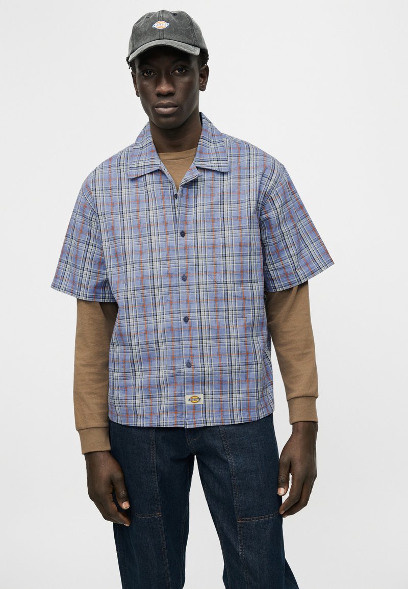 Giovane uomo con berretto grigio, camicia a maniche corte a quadri blu e arancioni sopra una maglia a maniche lunghe marrone, e jeans in denim scuro su sfondo neutro.