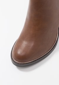 Stivale alla caviglia in finta pelle marrone con punta tonda, dettagli cuciti e una suola sottile di colore contrastante più scuro. Texture liscia con finitura lucida.