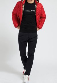 Rote gesteppte Jacke über einem schwarzen T-Shirt mit einem Logo, kombiniert mit schwarzen Hosen und weißen Sneakers mit roten Akzenten. Legere Kleidung.