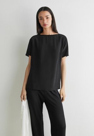 Giovane donna con lunghi capelli scuri che indossa una maglietta nera a maniche corte e pantaloni neri, tenendo un indumento bianco nella mano sinistra.