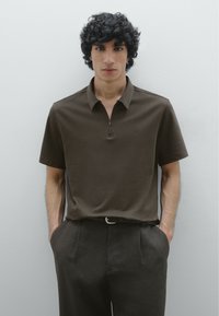 Polo shirt marrone con design a metà zip, maniche corte e una texture liscia, abbinato a pantaloni scuri e una cintura con una fibbia semplice.