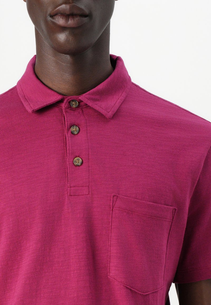 Polo magenta avec un col, trois boutons marron et une poche poitrine. Fabriqué en tissu texturé, montrant un léger éclat.