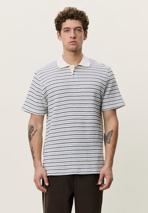 Ung mand med krøllet hår bærer en kortærmet, stribet strikket poloshirt og mørke bukser, der viser tatoveringer på begge underarme.