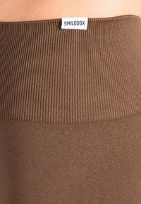 Bruine geribbelde tailleband met een gladde textuur, met een klein wit label met de tekst "SMILODOX" bevestigd aan de bovenrand.